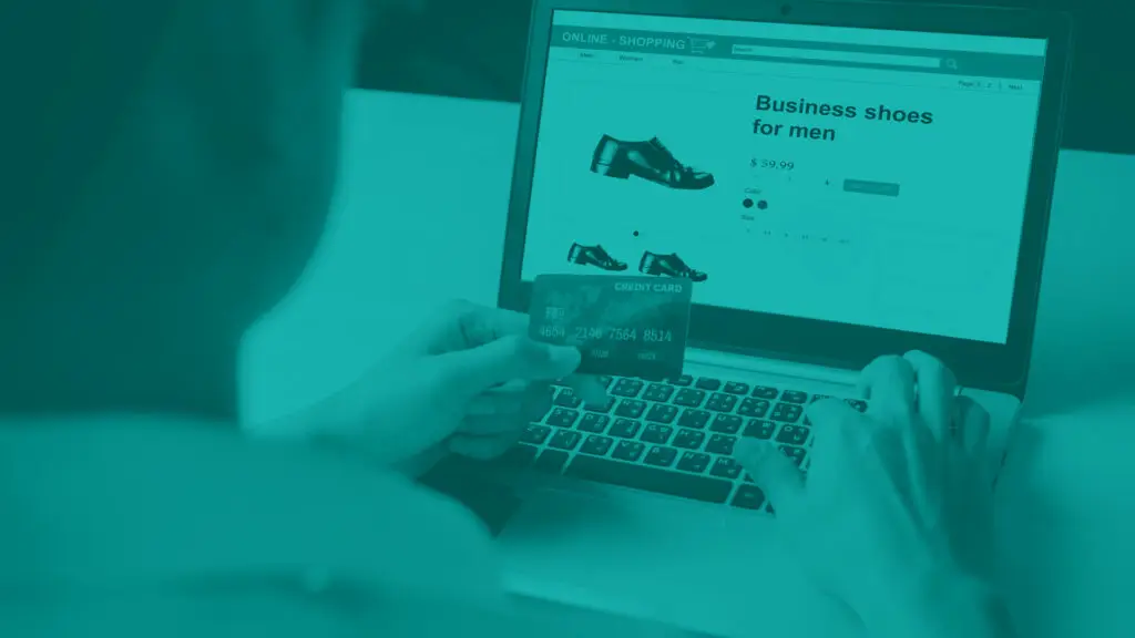 View from behind over a man's shoulder as he is shopping for shoes on a website using a laptop. He is holding a credit card in left hand ready to purchase.