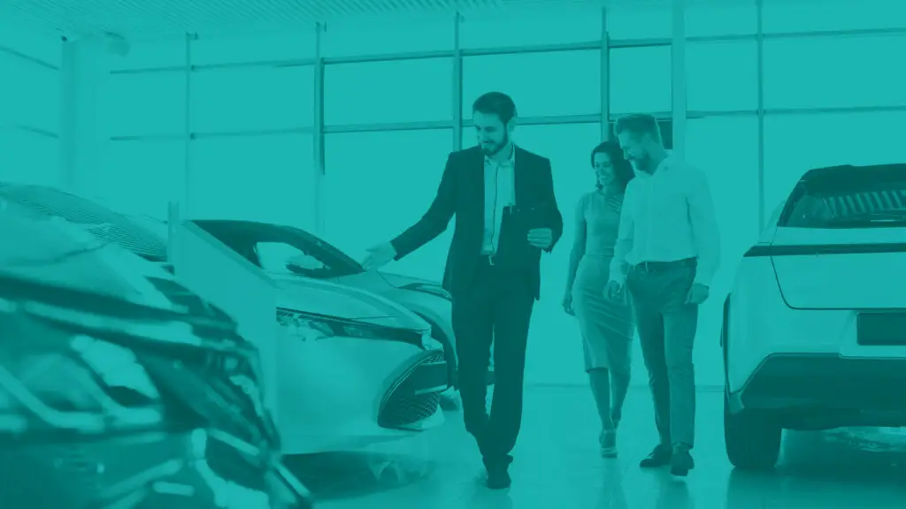 Automotive dealership salesman gesturing to new car in dealership showroom as a couple looks on shopping for a car